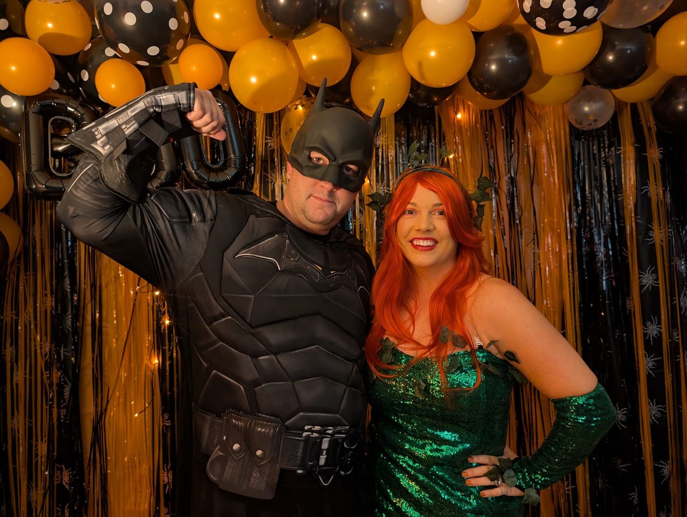 Batman flexing next to Poison Ivy under orange and black balloons