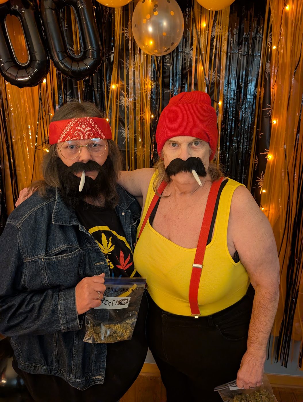 Two guests in bandanas, fake mustaches and suspenders in front of gold fringe